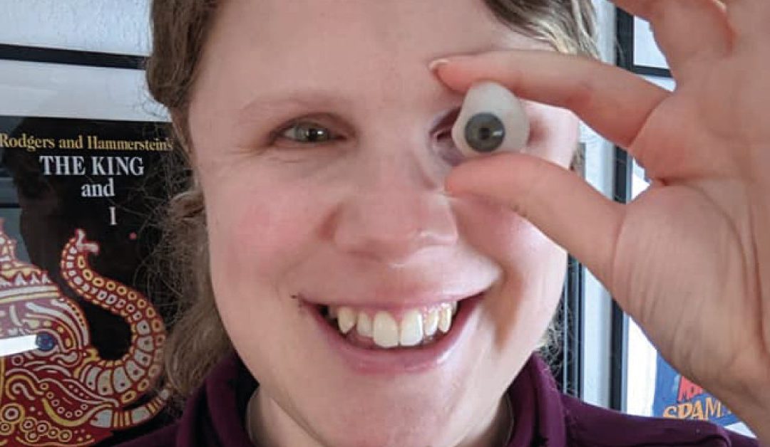 Nicole smiling at the camera whilst holding her prosthetic eye in front of her face