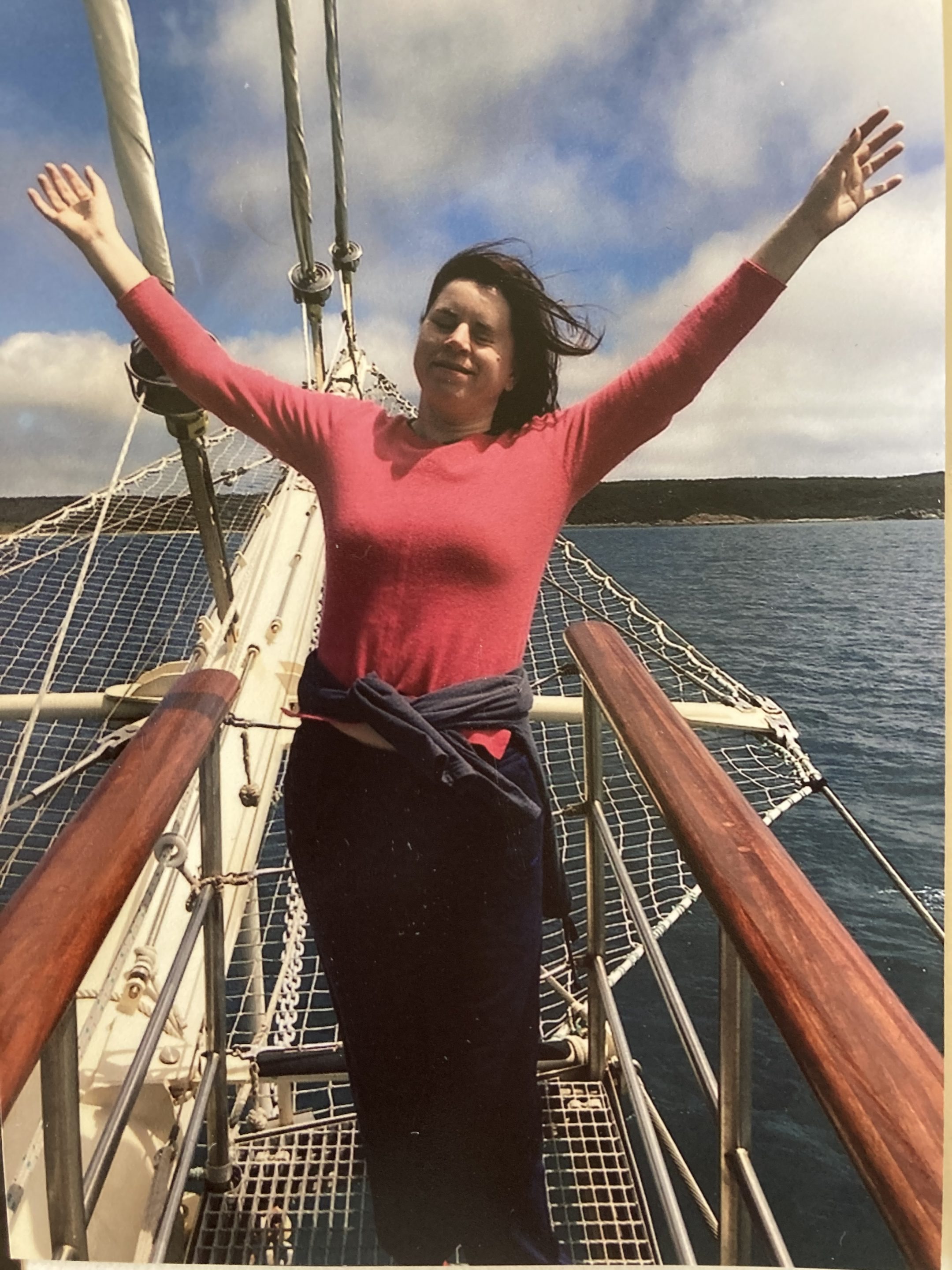 Ffion on a boat in the ocean, smiling and standing with her arms stretched outwards towards the sky.