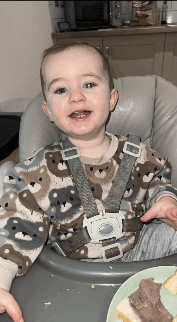 Kooper in a high chair, one eye has a white glow. He is smiling widely