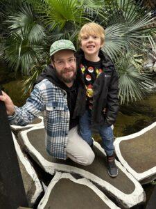Dad with his son, stood in an indoor rainforest with trees behind him