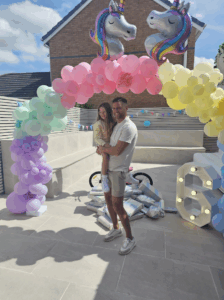 A dad is holding his daughter in front of a colourful balloon arch with unicorn balloons. There's a light up '6' decoration and unicorn balloons