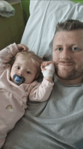 A dad and his daughter are lying next to each other, The daughter has her hand in a bandage and has a dummy in her mouth