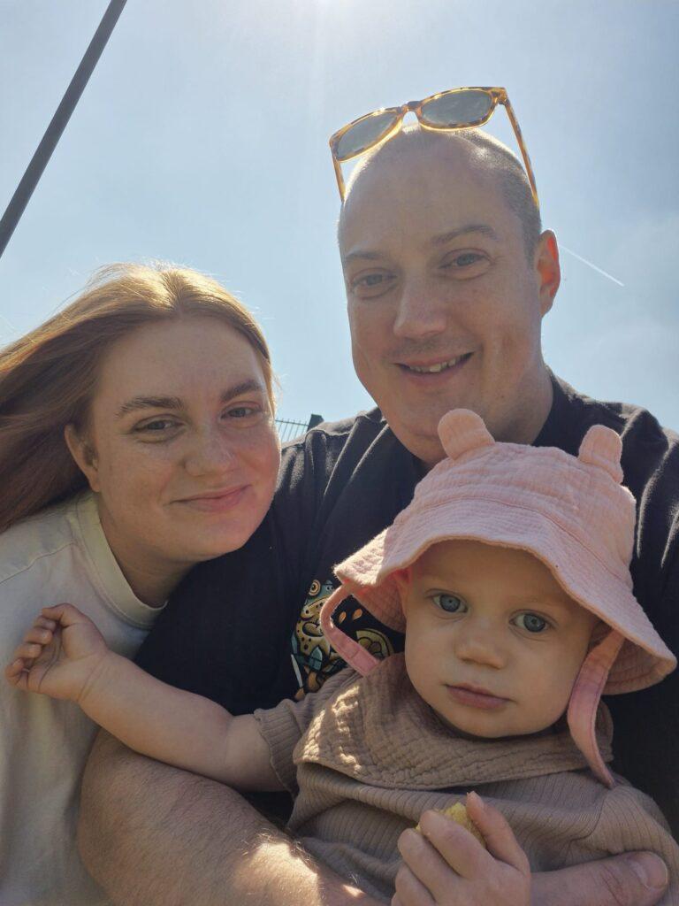 Raye wearing a pink hat, outside with her mum and dad in a selfie photo