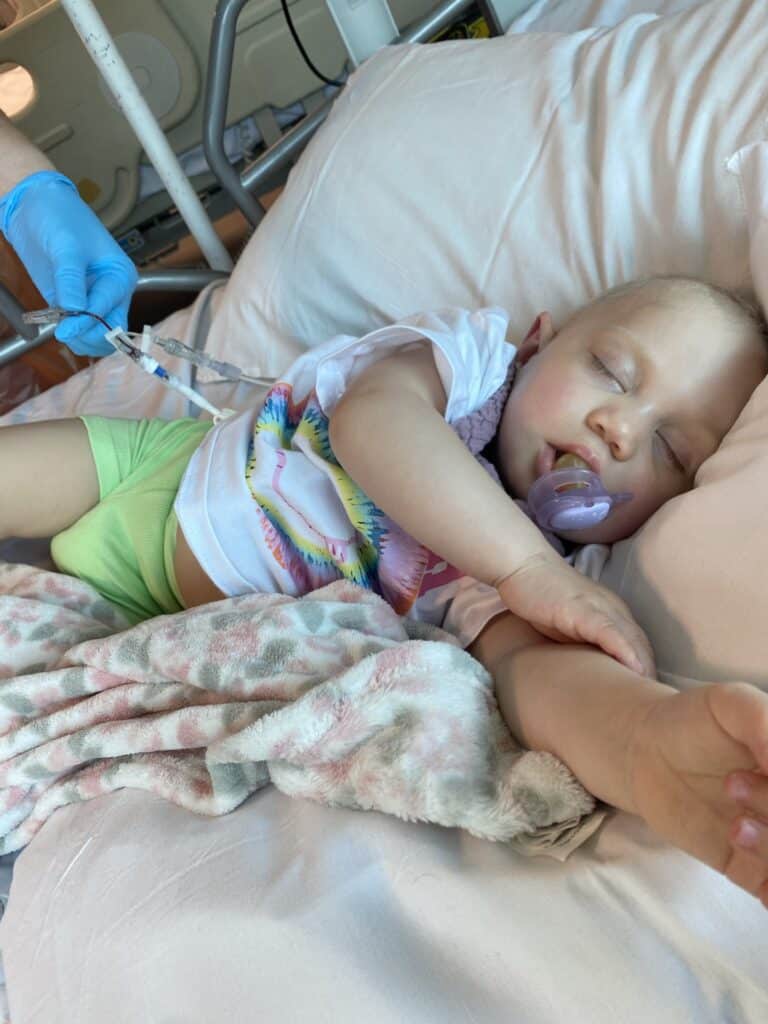 Raye lying on a hospital bed, asleep, with a dummy in her mouth